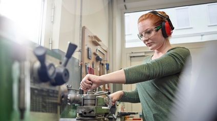Eine junge Frau mit Ohrenschutz und Schutzbrille arbeitet an der Werkbank
