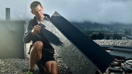 Ein Spengler auf einem Flachdach betrachtet ein Blechteil zur weiteren Verarbeitung. Im Hintergrund eine nebelige Landschaft