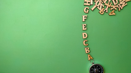 Ansicht von oben auf Holzbuchstaben in alphabetischer Reihung von A bis G von Taschenkompass zu Ansammlung weiterer Holzbuchstaben auf grünem Hintergrund verlaufend