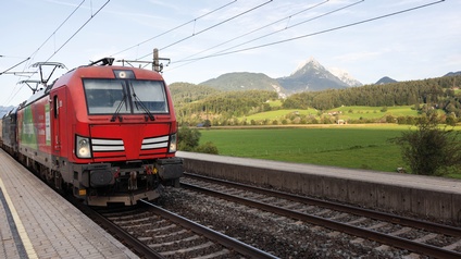 Rote Lok mit Güterwaggons fährt bei einem Bahnsteig ein, im Hintergrund zeigt sich eine ländliche Kulisse mit Wiesen, Wäldern und in einem Berg in der Ferne mit blauem Himmel