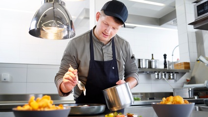 Person mit Cap und in Schürze steht in Edelstahlküche und tropft mit Löffel Flüssigkeit in eine Pfanne, hält in anderer Hand Topf, im Vordergrund verschwommen Schüsseln mit Eierschwammerl und lose Tomaten und Kräuter