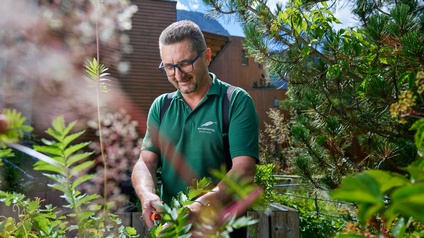 Lächelnde Person mit Brillen steht in Garten und schneidet Pflanzen, im Hintergrund verschwommen Holzfassade, ringsum grüne Pflanzen