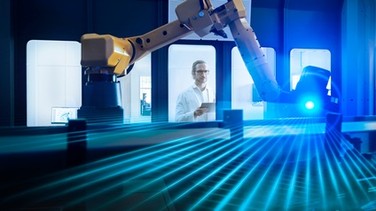 Person mit Brillen und in weißem Laborkittel blickt durch Glasscheibe auf Roboterarm mit blauem Laserlicht