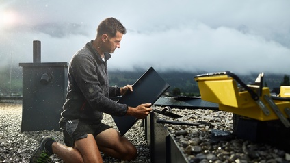 <br />
Ein Spengler auf einem Flachdach betrachtet ein Blechteil zur weiteren Verarbeitung. Im Hintergrund eine nebelige Landschaft