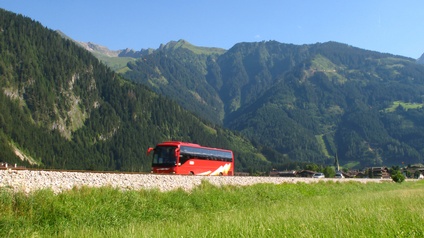 Ein roter Bus fährt durch eine Gebirgslandschaft, im Vordergrund eine grüne Wiese.
