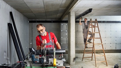 Ein Mann schneidet im Rohbau ein Abwasserrohr zurecht. Im Hintergrund wird es von einem Kollegen auf der Leiter an der Decke montiert.