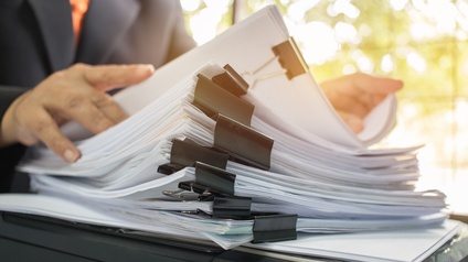 Vor einer Person steht auf einem Tisch ein Papierstapel. Die Person blättert durch den Stapel