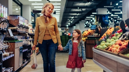 Eine erwachsene Person und ein Kind gehen Hand in Hand durch einen Supermarkt. Links von ihnen sind Behälter mit verschiedenen Nüssen. Rechts von ihnen sind Obstsorten. Die Person hält ein Einkaufsnetz mit Obst