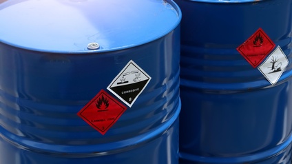 Zwei blaue Metallfässer mit roten quadratischen Stickern: Schwarzes Symbol einer Flamme, Schriftzug Flammable Liquid 3 und weiße quadratische Sticker mit Phiolen, die Flüssigkeit auf Hand & Baum tropfen sowie Fisch & blattloser Baum