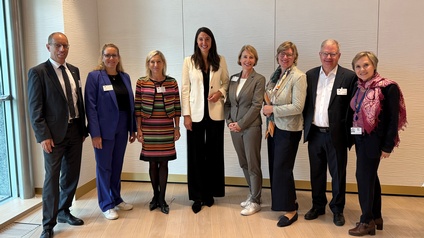 Gruppenfoto von mehreren Personen, die in einem Raum vor einer hellen Wand stehen. Die Personen tragen alle einen Ausweis um den Hals oder am Sakko und lächeln