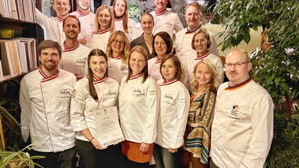 Gruppenfoto von 17 Personen. Fünfzehn Personen tragen Kochkleidung. Zwei Personen tragen Alltagskleidung. Drei Personen halten jeweils eine Urkunde