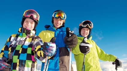 Kinder in Schikleidung mit Helm und Schibrille sowie Skistecken blicken freudig in die Kamera und halten die Hand mit Daumen nach oben, im Hintergrund zeigt sich eine winterliche Alpenlandschaft
