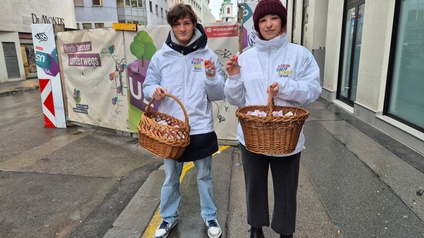 Porträt von zwei Personen, die auf einem Gehweg mit jeweils einem Einkaufskorb stehen. Im Einkaufskorb sind klein verpackte Schokokugeln. Beide Personen tragen weiße Jacken, auf denen mein Kaufstadt Wien steht