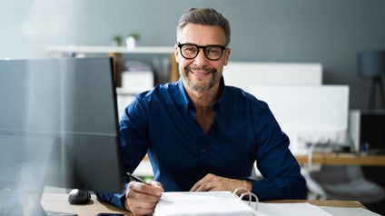 Porträt einer an einem Tisch sitzenden Person mit kurzen, grauen Haaren, Bart und Brille. Direkt vor der Person ist ein aufgeklappter mit einem Stapel Papier. Die Person hält einen Stift. Daneben ist ein Computer
