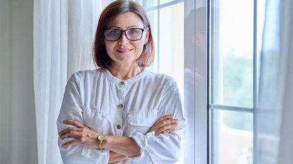 Eine Person steht mit verschränkten Händen vor einem Fenster mit einem Vorhang