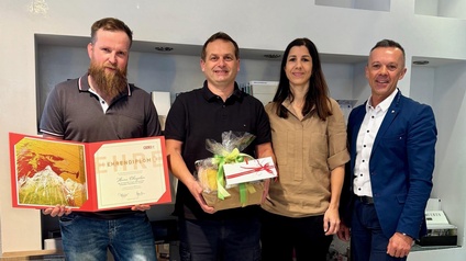 Vier Personen stehen nebeneinander in einem Innenraum vor einer Wand mit Firmenbeschilderung. Eine Person hält eine geöffnete Urkunde, eine weitere Person einen Geschenkkorb mit Schleife. Alle blicken in die Kamera und stehen dicht beieinander.