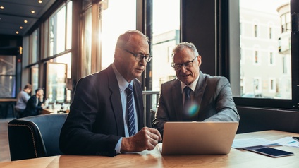 Zwei Personen in Anzügen sitzen an einem Tisch. Vor ihnen ist ein aufgeklappter Laptop, auf dessen Bildschirm beide blicken. Rechts neben dem Laptop sind Unterlagen. Im Hintergrund sitzen zwei weitere Personen an Tischen.
