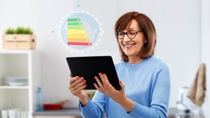 Person mit kurzen Haaren und Brille blickt freudig auf ein Tablet in einer Küche, daneben zeigt sich eine Grafik mit Energieeffizienzklassen