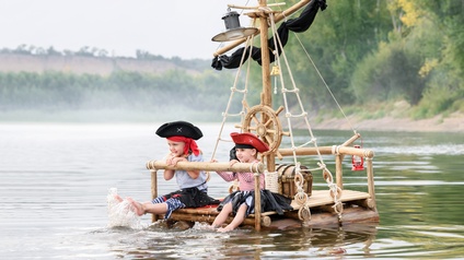 Verkleidete Kinder mit Piratenhut sitzen auf einem selbstgebauten Piratenfloß mit Schatztruhe und halten ihre Füße ins Wasser
