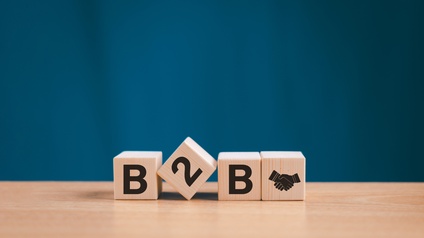 Auf einem Tisch aus hellem Holz sind vier Würfel platziert, wobei der zweite schief liegt, die anderen sind aufgestellt. Auf den Würfel steht B2B und am vierten Würfel ist das Symbol von sich schüttelnden Händen. Der Hintergrund ist blau