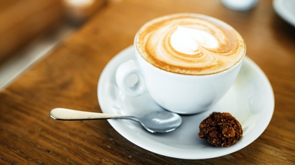 Nahaufnahme eines mit einem Milchschaumherz zubereiteten Cappuccino in einer weißen Kaffeetasse auf einem Holztisch. Neben der Kaffeetasse liegt auf dem Kaffeeteller ein kleiner Löffel sowie ein Keks.