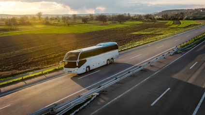 Seitliche Aufnahme eines weißen Autobusses auf einer zweispurigen Autobahn, in deren Mitte eine Abtrennung ist und danach wieder zwei Spuren sind. Im Hintergrund sind braune und grüne Felder