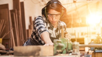 <br />
Person mit Ohrenschutz und Schutzbrillen hält Holzbalken auf Hobelbank, im Hintergrund verschwommen Holzbretter an Wand lehnend