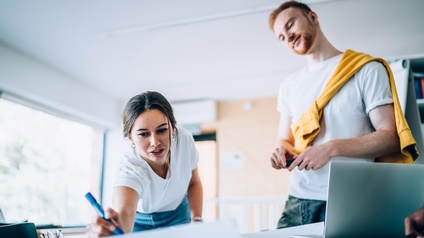 Person streckt sich über Tisch und markiert etwas mit Stift auf großem Papierbogen, andere Person sieht ihr mit geneigtem Kopf zu