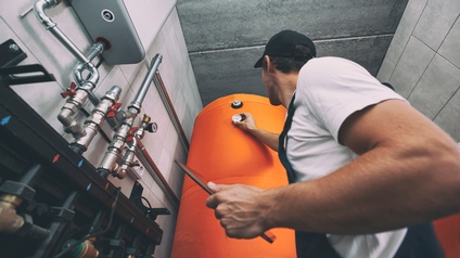 Person in Arbeitskleidung hält ein Tablet in der Hand und kontrolliert eine Messanzeige bei einem Wasserboiler in einem Heizsystem