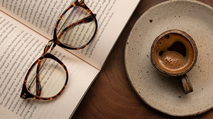 Gefüllte Kaffeetasse aus Steinzeug auf Unterteller stehend, daneben aufgeklapptes Buch mich Brille daraufliegend