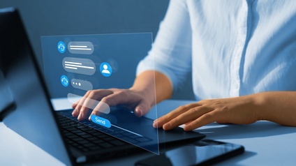 Nahaufnahme einer Person, die in einen Laptop tippt, der auf einer weißen Oberfläche steht. Daneben liegt ein Smartphone. Vor dem Bildschirm ist ein durchsichtiges Rechteck mit blauen Symbolen und Textfeldern. Man sieht das Symbol einer Person