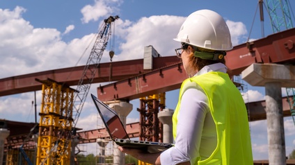 Eine Person ist von hinten fotografiert und hält einen Laptop in der Hand. Sie betrachtet die Metallkonstruktion einer Brücke, neben der sich auch ein Kran befindet. Am Himmel sind Wolken, es herrscht Sonnenschein.