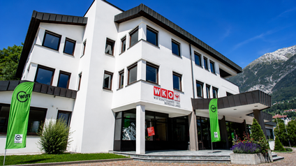 Außenansicht der Bezirksstelle Imst der Wirtschaftskammer Landeck. Das moderne, dreigeschossige Gebäude mit weißer Fassade und großen Fenstern trägt das WKO-Logo über dem Haupteingang. Vor dem Eingang stehen eine grüne WIFI-Fahne und eine Informationstafel der Wirtschaftskammer. Im Hintergrund sind Berge und ein klarer blauer Himmel zu sehen.