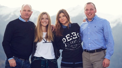 Vier Personen stehen nebeneinander im Freien vor einer Berglandschaft. Die Personen haben die Arme teilweise umeinander gelegt und blicken lächelnd in die Kamera. Sie tragen Freizeitkleidung.