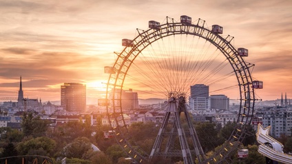 Großes Riesenrad im Wiener Prater mit roten Gondeln im Licht des Sonnenuntergangs, ringsum Baumkronen und im Hintergrund Gebäude der Stadt