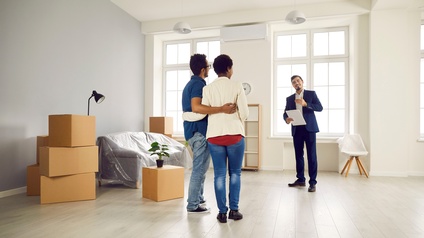 Rückenansicht zweier Personen, die eng nebeneinander und die Arme gegenseitig am Rücken liegend, in einem Wohnraum stehen. Ihnen gegenüber steht eine weiter Person mit einem Tablet. Links neben ihnen stehen Kartons