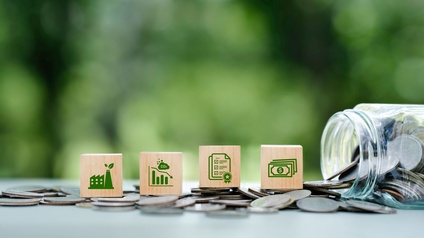 Holzwürfel mit Symbolen für Industrie, CO₂-Reduktion, Zertifizierung und finanzielle Förderung stehen auf verstreuten Münzen. Rechts liegt ein umgekipptes Glas mit herausgerollten Münzen. Der unscharfe grüne Hintergrund vermittelt einen Bezug zu Nachhaltigkeit, Umwelt- und Klimaschutzmaßnahmen in Unternehmen.