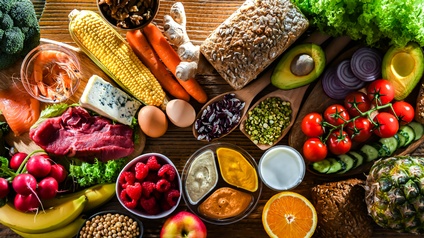 Topshot verschiedener Lebensmittel auf einer hölzernen Unterfläche, zum Beispiel Brot, Eier, Käse, Fleisch, Lachs, Bananen, Tomaten, Aufstriche, Samen, Avocados, Radieschen, Joghurt, Karotten, Ingwerwurzel