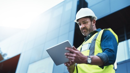 Eine Person in Nahaufnahme schaut auf ihr Tablet. Im Hintergrund ist ein Hochhaus mit einer Glasfront.