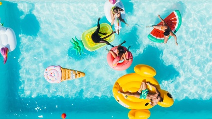 Top-Shot eines blauen Wassers. Auf dem Wasser sind verschiedene, große Plastikobjekte in Formen von einer Wassermelone oder einer Eistüte. Auf manchen Plastikobjekten sind Personen in Badebekleidung.