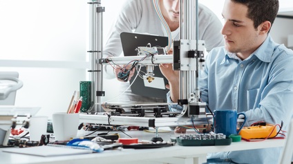 Personen arbeiten an einem 3D Drucker, am Tisch sind verschiedene technische Komponenten ersichtlich