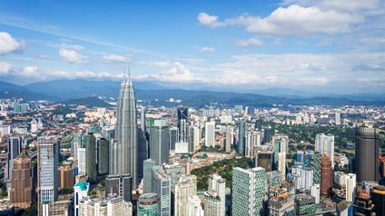Blick von oben auf die modernen Gebäude von Kuala Lumpur, im Hintergrund erkennt man eine bewaldete hügelige Landschaft.