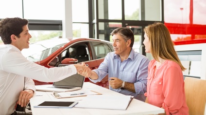 Drei Personen sitzen an Tisch, zwei reichen sich die Hand, im Hintergrund rotes Auto