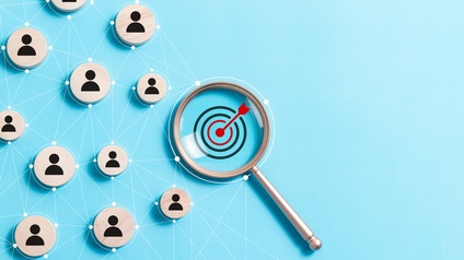 Auf hellblauem Untergrund sind  Kreise aus hellem Holz platziert, auf denen das Symbol  einer Person ist. Die Kreise sind durch Linien, die von Punkten ausgehen, verbunden. Daneben ist eine Lupe, die das Symbol einer Zielscheibe mit rotem Pfeil fokussiert