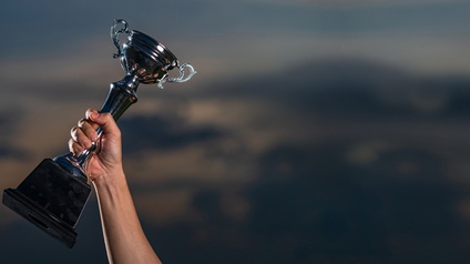 Ein Arm hält einen silbernen Pokal in die Höhe, im Hintergrund ein dunkler, bewölkter Himmel