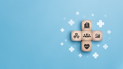 Auf hellblauem Hintergrund liegen vier Holzwürfel, die ein Kreuz bilden. Auf jedem Würfel ist ein anderes Symbol, zum Beispiel ein Krankenhaus oder ein Herz mit einer Frequenz darin