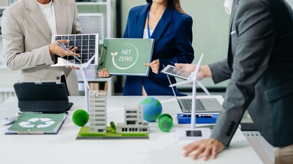 Mehrere Personen im Ausschnitt, nur Oberkörper und Hände ersichtlich, an Tisch stehend mit Modellen wie das eines Windrades, eine Person hält Schild mit Aufschrift Net Zero in Händen
