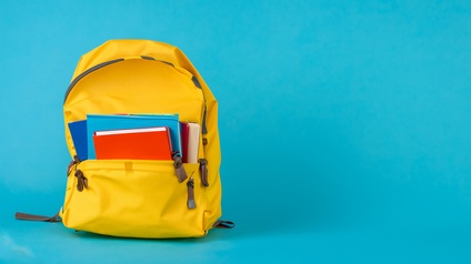 Ein gelber Rucksack steht auf einem blauen Hintergrund vor einer blauen Wand in der linken Hälfte. Ein Fach des Rucksacks ist geöffnet. Aus dem Fach schauen die Hälften von Notizbüchern, Büchern und Heften