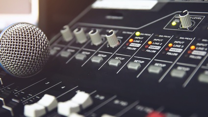Nahaufnahme eines Mischpults, auf dem ein paar Leuchtkontrollen orange oder gelb aufleuchten. Auf dem Mischpult liegt ein Mikrofon