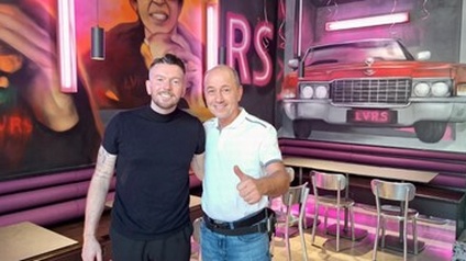 Porträt von zwei Personen, die dicht nebeneinander in einem Lokal stehen. Die rechte Person zeigt den Daume nach oben